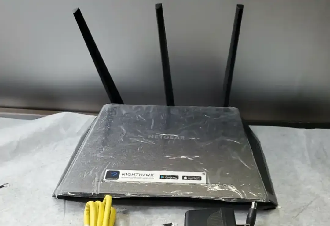 A Netgear Nighthawk AC1900 router with three antennas raised, still wrapped in plastic, beside a yellow ethernet cable and power adapter